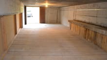 This image shows the interior of a garage or storage space. The space has concrete walls and a wooden floor. There is a window at the end of the room, allowing natural light to enter. The room appears to be empty and unfinished, with a simple light fixtur