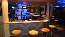 Modern bar with a sleek, geometric design. The bar counter is made of wood and has a granite-like surface. The back bar is well-stocked with various liquor bottles and glassware. There are yellow bar stools in front of the counter, and the lighting create