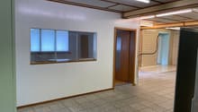 The image shows a large open office space with a tiled floor, white walls, and wooden beams on the ceiling. There is a window with blinds and a doorway leading to another room. The space appears to be a commercial or office setting.