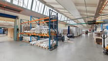 Large industrial warehouse with high ceilings, storage racks, and various materials and equipment stored on the premises. The space appears to be used for storage and distribution of goods.