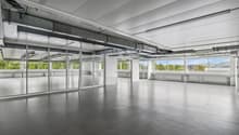 Large open office space with high ceilings, concrete floors, and floor-to-ceiling windows providing natural light and views of the surrounding greenery. The space has a modern, minimalist design with exposed ceiling beams and ductwork.