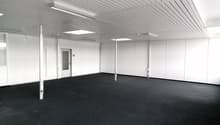 Large open office space with white walls, gray ceiling panels, and black carpeted floor. The room appears to be empty and ready for use.