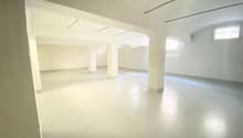 Large open space with white walls, concrete floor, and several windows providing natural light. The room appears to be an empty, unfinished space, possibly a loft or studio apartment.
