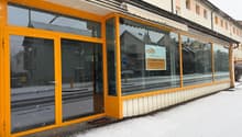 Exterior of a commercial building with glass windows, double-door entrance, signage, and orange frames. Snow on the ground and street.