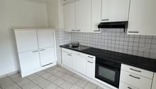 Spacious kitchen with white cabinets, black countertops, tiled backsplash, and built-in appliances including a dishwasher. The floor is covered with gray square tiles.
