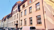 Multi-story building with beige walls, red shutters, many windows, ground floor and upper floors with balconies, white van parked in front.