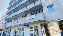 A multistory residential building with balconies, glass doors and windows, and signage indicating stores or offices on the ground floor.