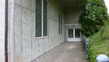Concrete building with two large windows, brick pathway, white entrance door, and lamp on the right