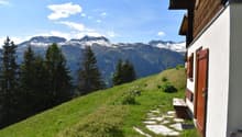 A wooden chalet situated on a grassy hillside with a stunning view of snow-capped mountains in the background. The chalet has wooden shutters and is surrounded by pine trees.