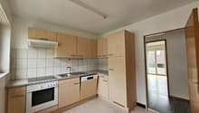 A kitchen with a gas stove, oven, sink, dishwasher, and countertop. Wooden cabinets and countertops are on the walls, and a power outlet is mounted on the wall. There is an entrance to the left side, and a door with a metal handle is open.