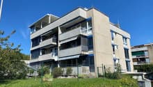 multi-storey building, concrete facade, balconies on upper floors, green fencing surrounding