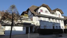 Multi-story residential building, white walls, brown roof, multiple balconies, ground floor entrance, staircase, hedge, large tree, small bench