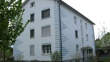 3-story white building, windows with shutters, chimney, entry area with canopy, greenery and plants surrounding the building