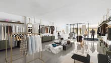 Modern store with glass doors, shelves for clothing, and golden poles holding clothing. Inside has couches, tables, and displays for clothing and accessories.