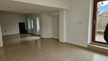 The image shows a large open room with a tiled floor. The walls are painted in a neutral color and there is a window overlooking what appears to be a garden or outdoor area. The room has a simple, minimalist design with no visible furniture or decor.