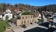 A modern multi-story building with a parking lot in the foreground, surrounded by a residential neighborhood with traditional houses and buildings in a hilly, forested area.