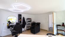 Spacious hair salon with modern decor, including black salon chairs, shelves with hair products, and a large circular mirror. The room has a bright, open layout with recessed lighting fixtures on the ceiling.