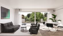 Spacious living room with large windows overlooking a lush, green outdoor area. The room features a grey sofa, black armchairs, a white dining table with chairs, and a potted plant. The walls are white, and the floor is made of wood. There is also a frame