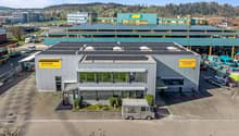 Large commercial building with solar panels on the roof, yellow and blue accents, parking lot in front, delivery van parked in the lot