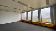Large open office space with floor-to-ceiling windows providing a view of the surrounding cityscape. The room has a gray carpeted floor and a suspended ceiling with recessed lighting. There are built-in benches along the windows.