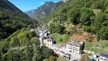 Old building located in a mountainous region with lush greenery, several vehicles parked outside, surrounded by other buildings.