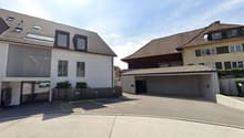 A multi-story residential building with a garage and parking spaces in the foreground. The building has a mix of modern and traditional architectural styles, with a combination of flat and sloped roofs. There are several windows and balconies visible on t