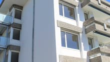 Multi-story residential building, white exterior, balconies on upper floors, large windows, modern architecture, grey stone accents, flat roof with ridge tiles