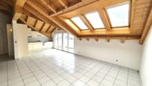 Spacious open-plan living area with vaulted wooden ceiling, large windows, and sliding glass doors leading to a balcony or terrace. The kitchen is located in the corner with white cabinets and tiled flooring throughout the space.