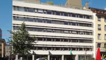 Modern white building with glass windows, rooftop terrace, plants in front, parked car