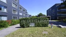 Modern apartment building with blue exterior, multiple windows, and a sign reading 'IM MITTELDORF'. The building is surrounded by greenery and has a driveway.