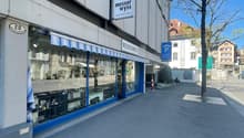 Commercial building with a shopfront, blue awning, and a sign reading 'Messer Wyss'. The building has a modern design with a white exterior and blue accents. The shopfront features large glass windows displaying items inside. There is a parking sign and a