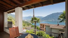 Wooden balcony with a scenic view of a lake surrounded by mountains and palm trees. The balcony has a wooden chair and a straw hat, suggesting a relaxing outdoor space.