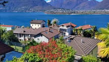 Scenic view of a lakeside town with colorful houses, palm trees, and mountains in the background. The houses have red tile roofs and are surrounded by lush greenery.