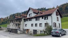 Large building with multiple floors, tiled brown roof, white walls, several windows, located in a scenic setting with green fields and trees