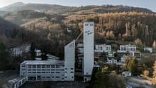 An industrial building with a tall shaft named Senn fullter. It is surrounded by trees and mountains. There are several houses and buildings nearby, and a car is parked on the road.