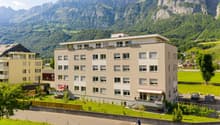 Multi-story apartment building with balconies, surrounded by green landscaping and with a view of the mountains in the background. A person riding a scooter is visible in the foreground.