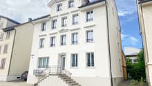 White building with multiple windows, stairs leading to the entrance, car parked in front, small garden with plants