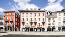 This image shows a row of multi-story buildings with arched entrances and balconies in a European-style town square. The buildings have a mix of orange, pink, and white facades, and there is a Coop supermarket visible on the ground floor of one of the bui