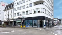 High-rise building, glass windows, black and white facade, store fronts, cafe with chairs, man walking on the sidewalk, greenery in front