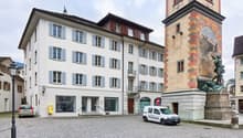 This image shows a historic town square with a tall clock tower and several multi-story buildings with traditional Swiss architectural styles. The buildings have white facades with blue shutters and balconies. There is a cobblestone plaza in the foregroun