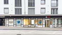 The image shows the exterior of a UBS bank branch. The building has a white facade with horizontal striped windows. The entrance is located in the center, with the UBS logo displayed prominently. There are several glass doors and windows, allowing visibil