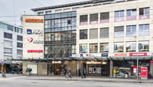 Multi-story commercial building with various retail stores and businesses, including Migros, Axa, Akros, and Denner. The building has a modern glass and concrete facade with signage for the different companies. There are parking spaces in front of the bui