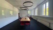 Empty conference room with white walls, black carpet, glass whiteboards, and wooden long table. Large windows with white frames and red chairs.