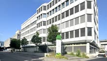 Multi-story office building with a modern, angular design featuring large windows and louvered facades. The building has a prominent green sign labeled 'Block 22' on the ground floor. There are trees and landscaping around the building, and a paved road i