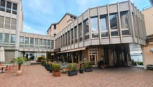 Modern building with glass windows, outdoor seating area, potted plants, brick pavement, and a view of the sea.