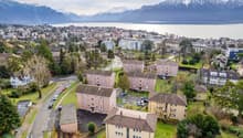 Aerial view of apartment buildings, some with parking areas, set in a picturesque location with mountains and a lake in the background.