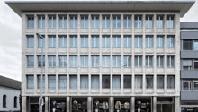 Concrete building, large windows, multiple floors, modern architecture, commercial facade, shopfronts, street-level entrance, grey exterior