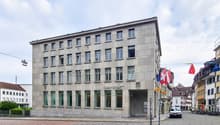 3 story building, concrete exterior, many windows, entrance at street level, flag at entrance