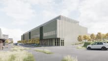 A large modern industrial or commercial building with a rectangular, multi-story design featuring a mix of concrete and glass materials. The building has a sleek, contemporary architectural style with vertical lines and a prominent entrance. The surroundi