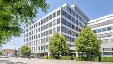Raised ground floor, office building, several floors, many windows, hedges and trees in front, street with parked car